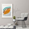 Framed contemporary poster design of the lens in teal, brown, orange, and gray. The painting is hanging on a gray wall over light gray chair and cactus.