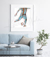 Framed watercolor painting of a soccer player kicking a ball. Painting shows a soccer player at a muscular and skeletal level. The painting is hanging over a blue couch.