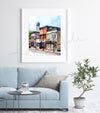 Framed watercolor painting of buildings on Main Street in Waunakee, Wisconsin. The painting is hanging over a blue couch.